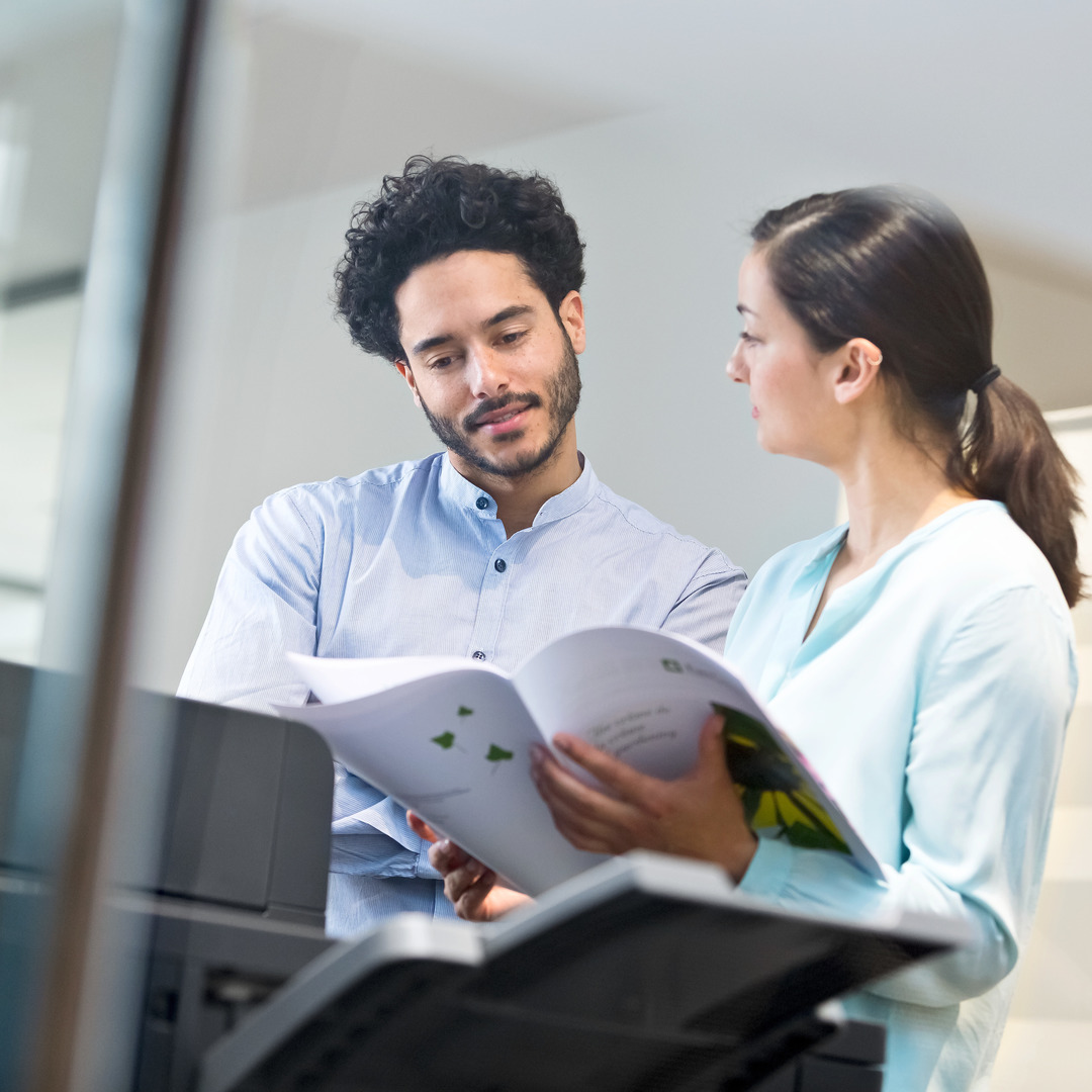 Two colleagues reviewing printed documents beside a multifunction printer in a modern office